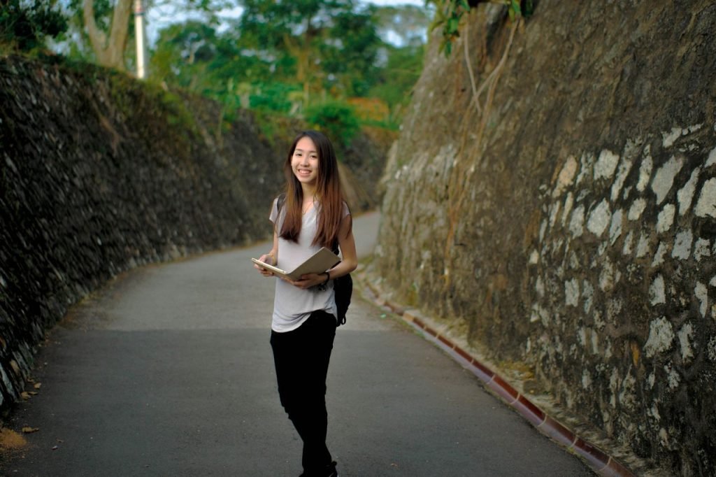 Asian woman smiling while holding books on a stone pathway outdoors.