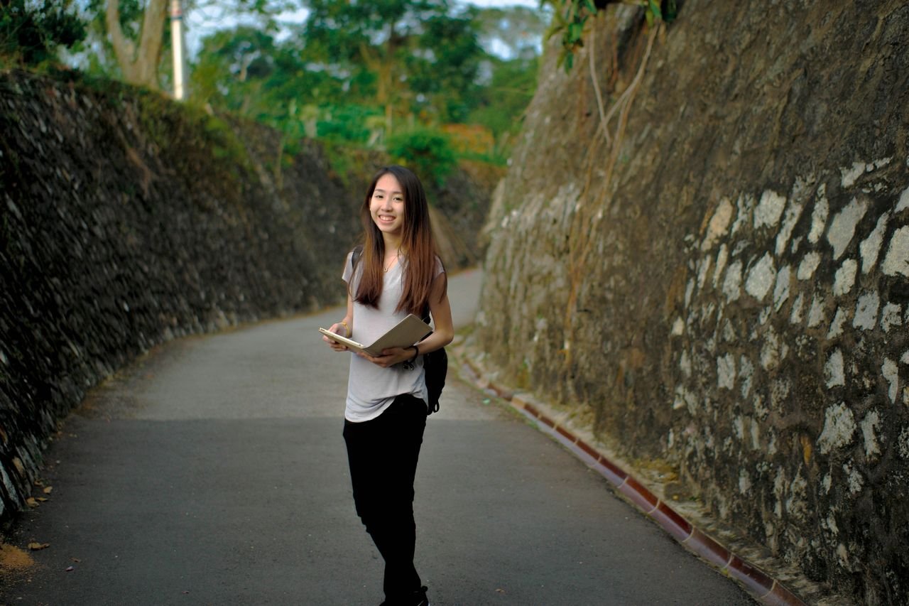 Asian woman smiling while holding books on a stone pathway outdoors.